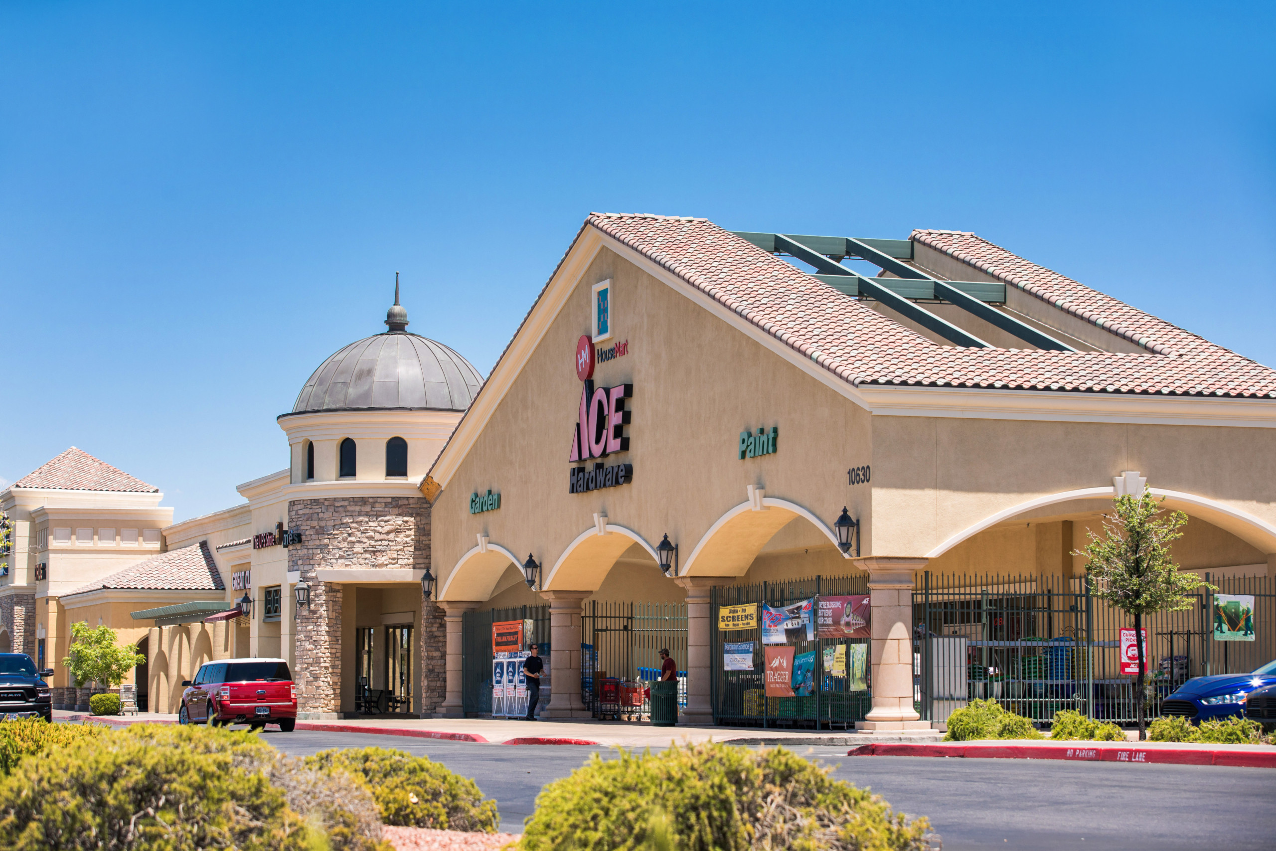 Commercial retail shopping center with storefronts and parking area in sunny weather.